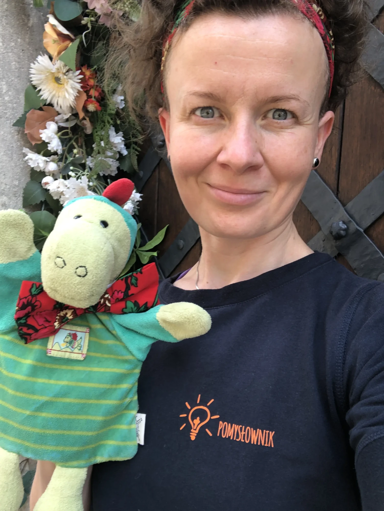 Zdjęcie potretowe kobiety, kobieta ma ciemne włosy, założoną czarną koszulkę z napisem POMYSŁOWNIK, a w ręce trzyma maskotkę Smoka Wawelskiego.