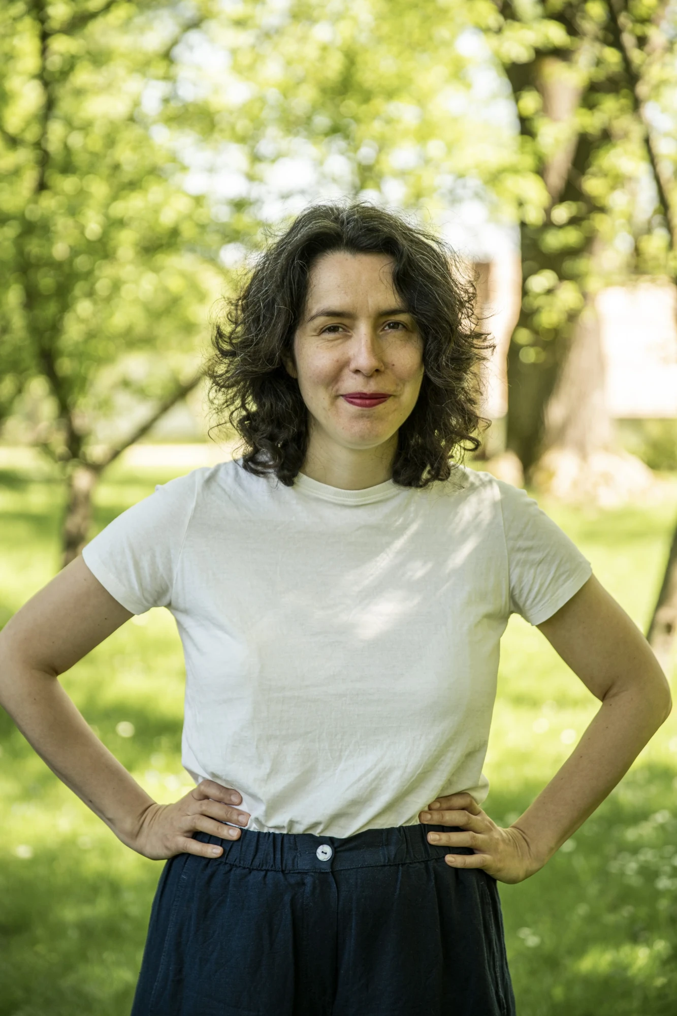 Ciemnowłosa kobieta pozuje do zdjęcia, ma ręce na biodrach, uśmiecha się. W tle widać drzewa.