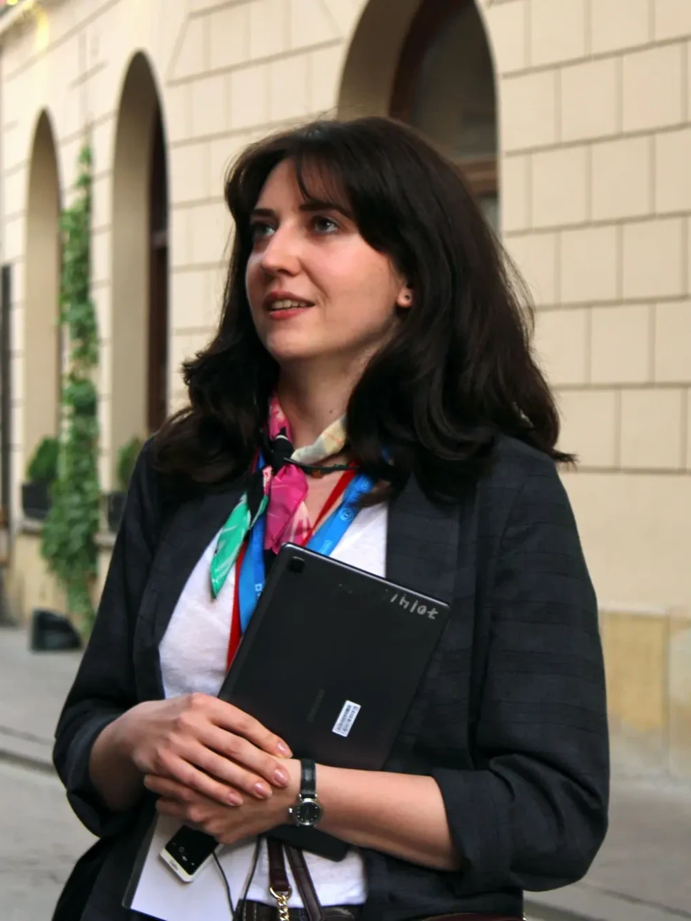 Ciemnowłosa kobieta, trzymająca w rękach tablet, mowi do ludzi.