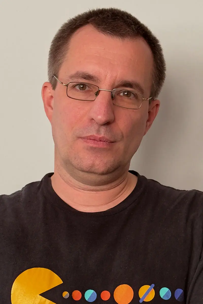 Zdjęcie portretowe mężczyzny. Mężczyzna ma ciemne, krókie włosy, okulary z cienkimi srebrnymi oprawkami oraz założoną koszulkę z nadrukiem.