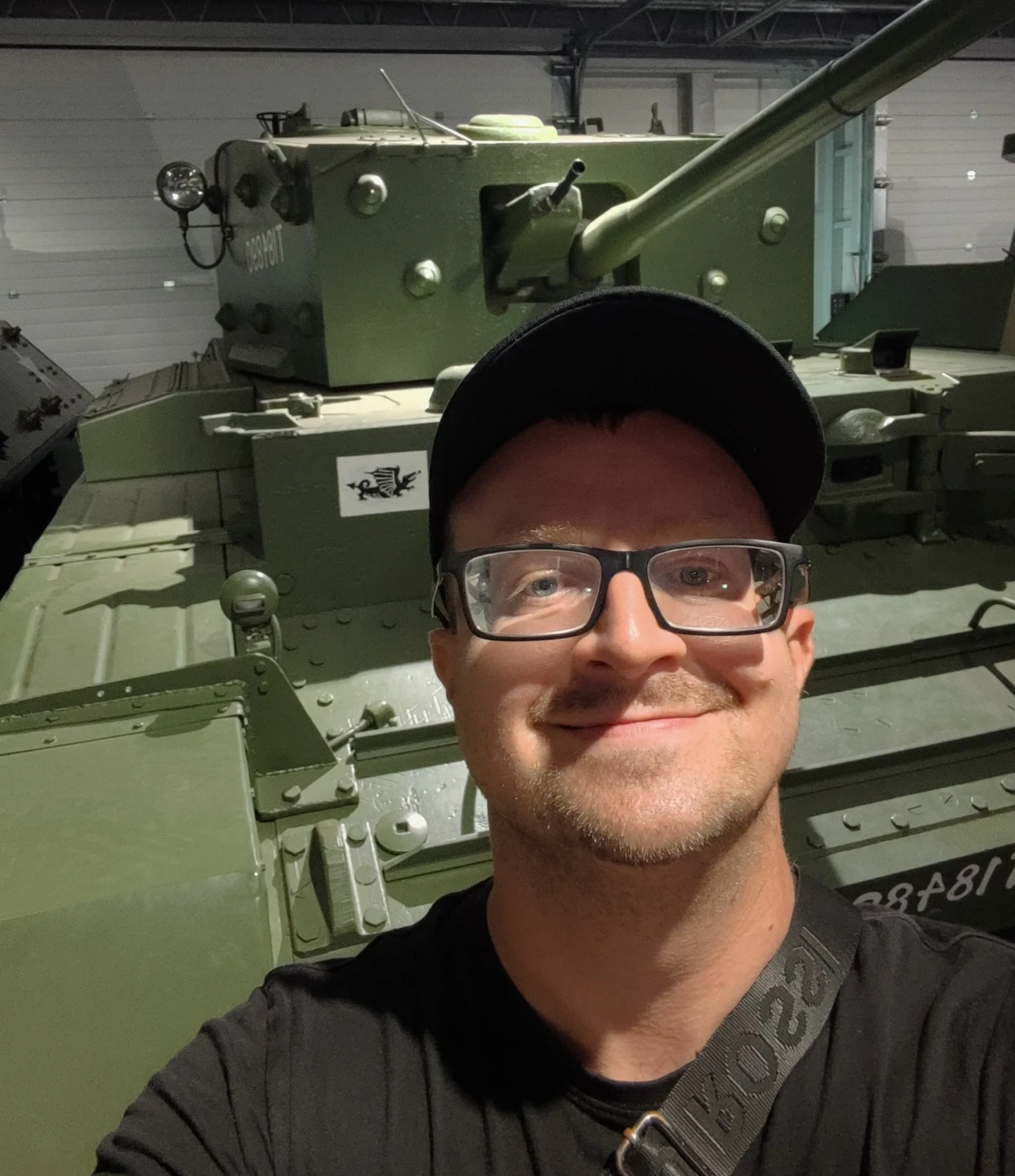 Mężczyzna w okularach i czapce z daszkiem uśmiecha się do zdjęcia, w tle czołg.