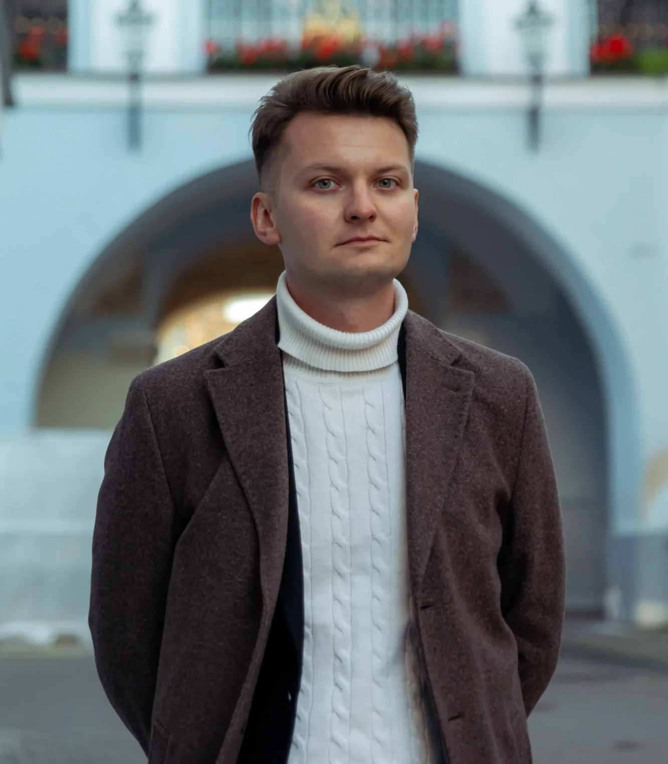 Zdjęcie portretowe młodego mężczyzny. Mężczyzna ma krótkie jasnobrązowe włosy, założony biały swter i brązową marynarkę. W tle widać krakowską ulicę.