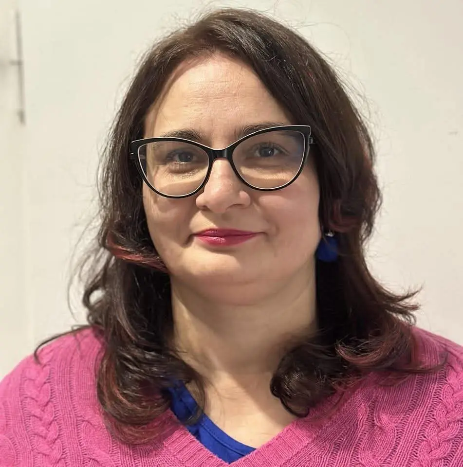 Zdjęcie potretowe kobiety, kobieta ma brązowe włosy, pomalowane na różowo usta, niebieskie kolczyki, różowy sweter i okulary z czarnymi oprawkami.